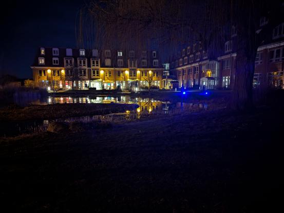 Nächtliche Aufnahme eines großen Hotelgebäudes aus roten Backsteinen mit mehreren Stockwerken. Die warm erleuchteten Fenster und Außenlampen spiegeln sich in einem kleinen Teich der vorgelagerten Parkanlage. Rechts ragt eine kahle Trauerweide ins Bild. Vor dem Hotel leuchten zwei auffällige blaue Lichter. Der dunkle Nachthimmel und das dunkle Gras im Vordergrund rahmen die stimmungsvolle Szene ein.